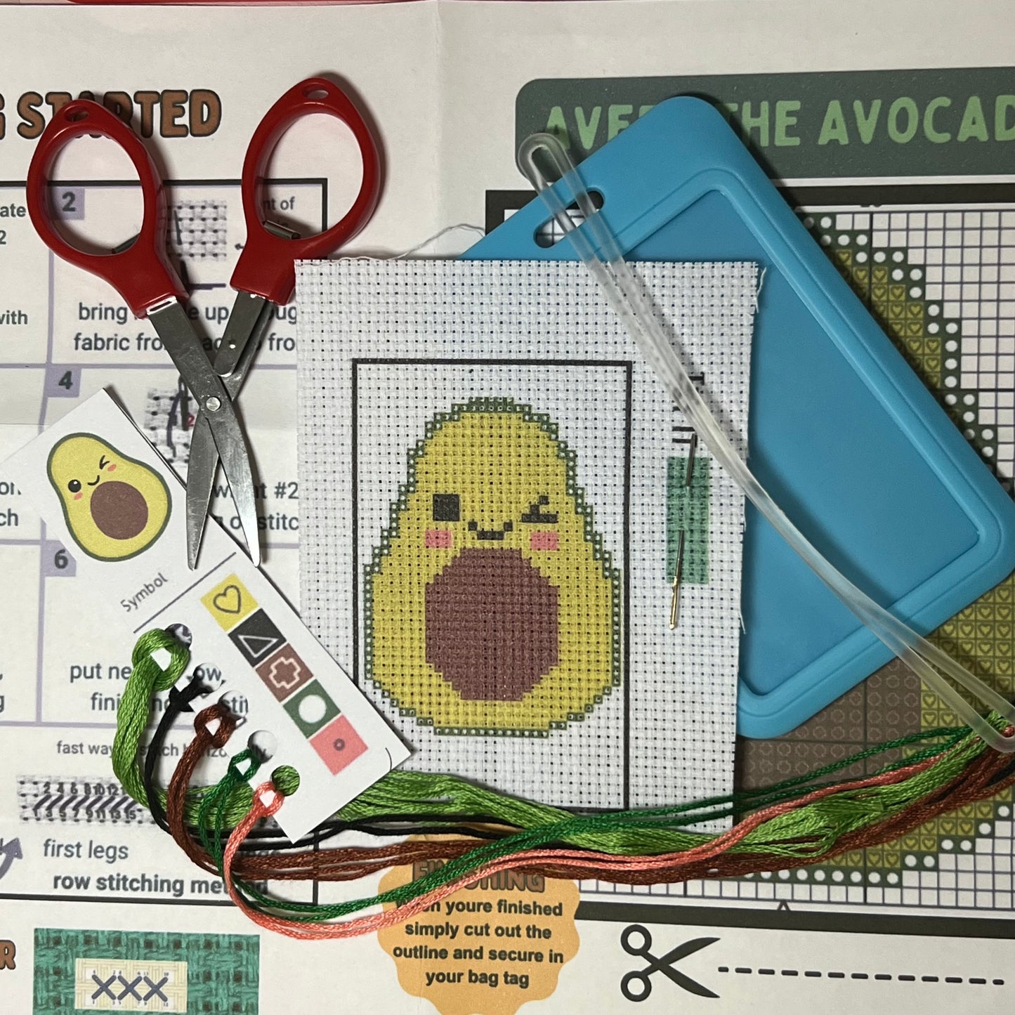 Avery the Avocado mini cross stitch DIY luggage tag kit with a light blue bag tag, luggage tag attachment, Scissors, needle and thread, stamped Aida fabric, color instructions