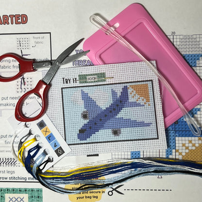 Blue Airplane in the Clouds mini cross stitch DIY luggage tag kit with a pink bag tag, luggage tag attachment, Scissors, needle and thread, stamped Aida fabric, color instructions