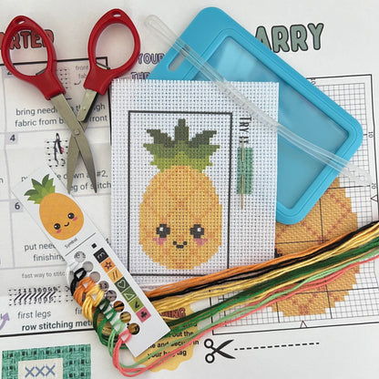 Larry the Pineapple mini cross stitch DIY luggage tag kit with a light blue bag tag, luggage tag attachment, Scissors, needle and thread, stamped Aida fabric, color instructions