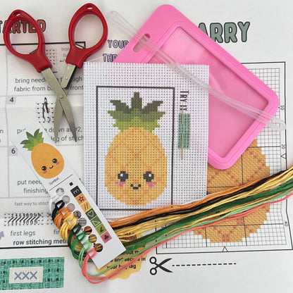 Larry the Pineapple mini cross stitch DIY luggage tag kit with a pink bag tag, luggage tag attachment, Scissors, needle and thread, stamped Aida fabric, color instructions