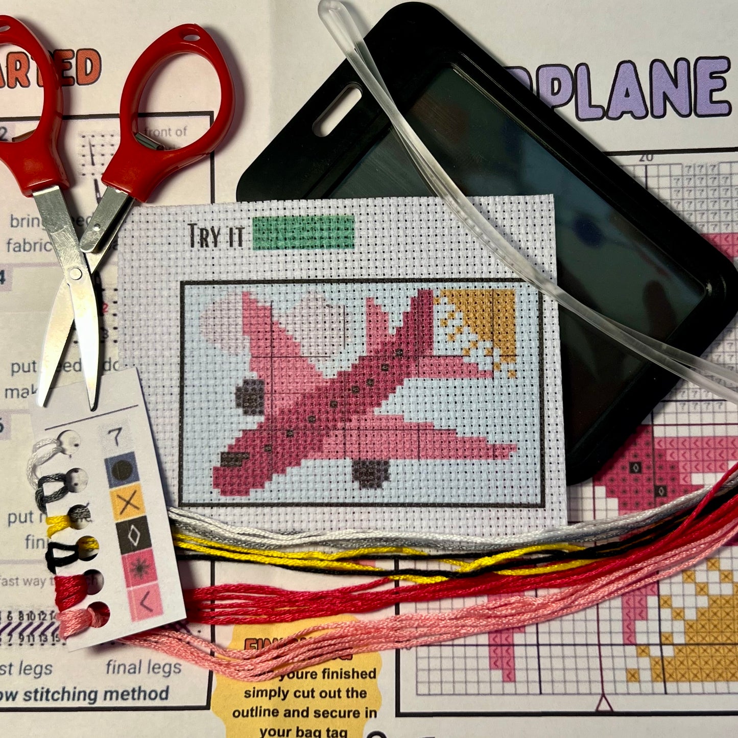 Pink Airplane cross stitch DIY luggage tag kit with a black bag tag, luggage tag attachment, Scissors, needle and thread, stamped Aida fabric, color instructions