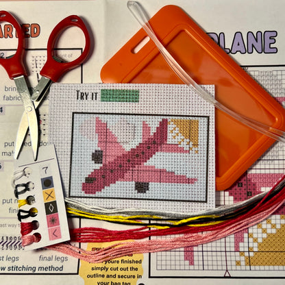 Pink Airplane cross stitch DIY luggage tag kit with a orange bag tag, luggage tag attachment, Scissors, needle and thread, stamped Aida fabric, color instructions